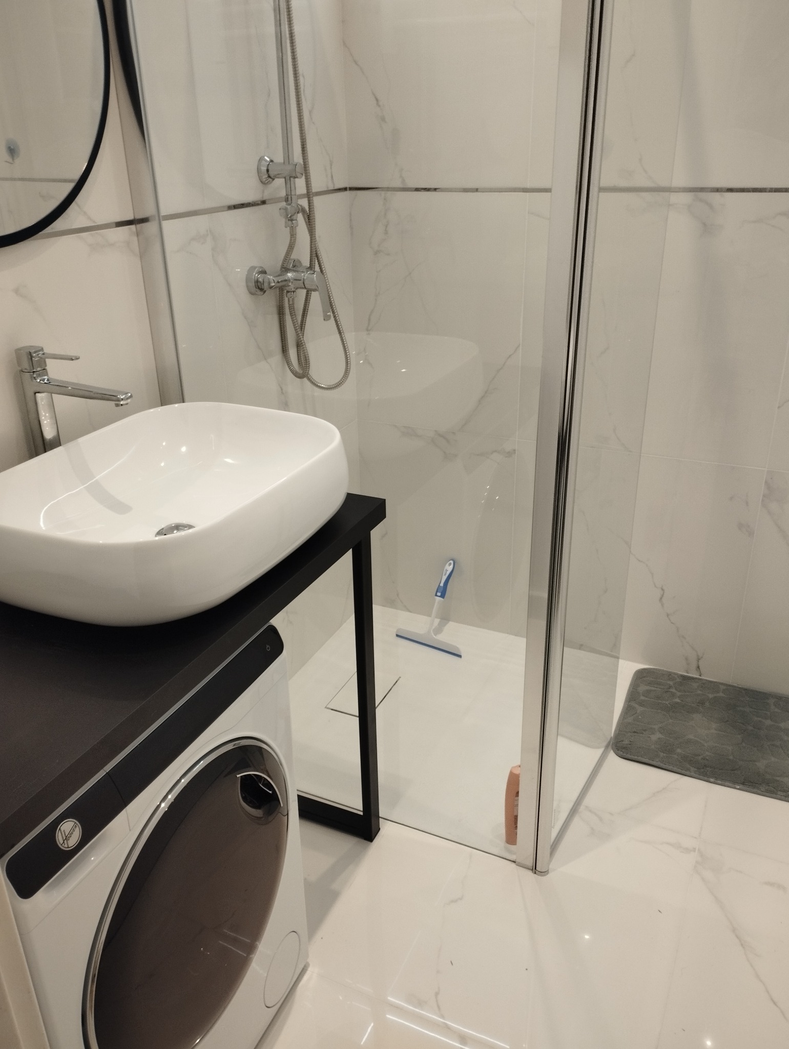 Nowoczesna łazienka z kabiną prysznicową typu walk-in, umywalką nablatową na czarnym blacie i pralką. Białe płytki imitujące marmur.