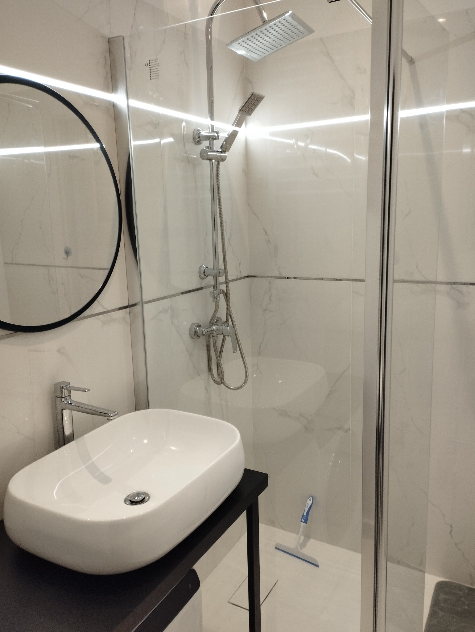 Nowoczesna łazienka z owalną umywalką na czarnym blacie, okrągłym lustrem i kabiną prysznicową z deszczownicą. Białe marmurowe płytki na ścianach i podłodze.