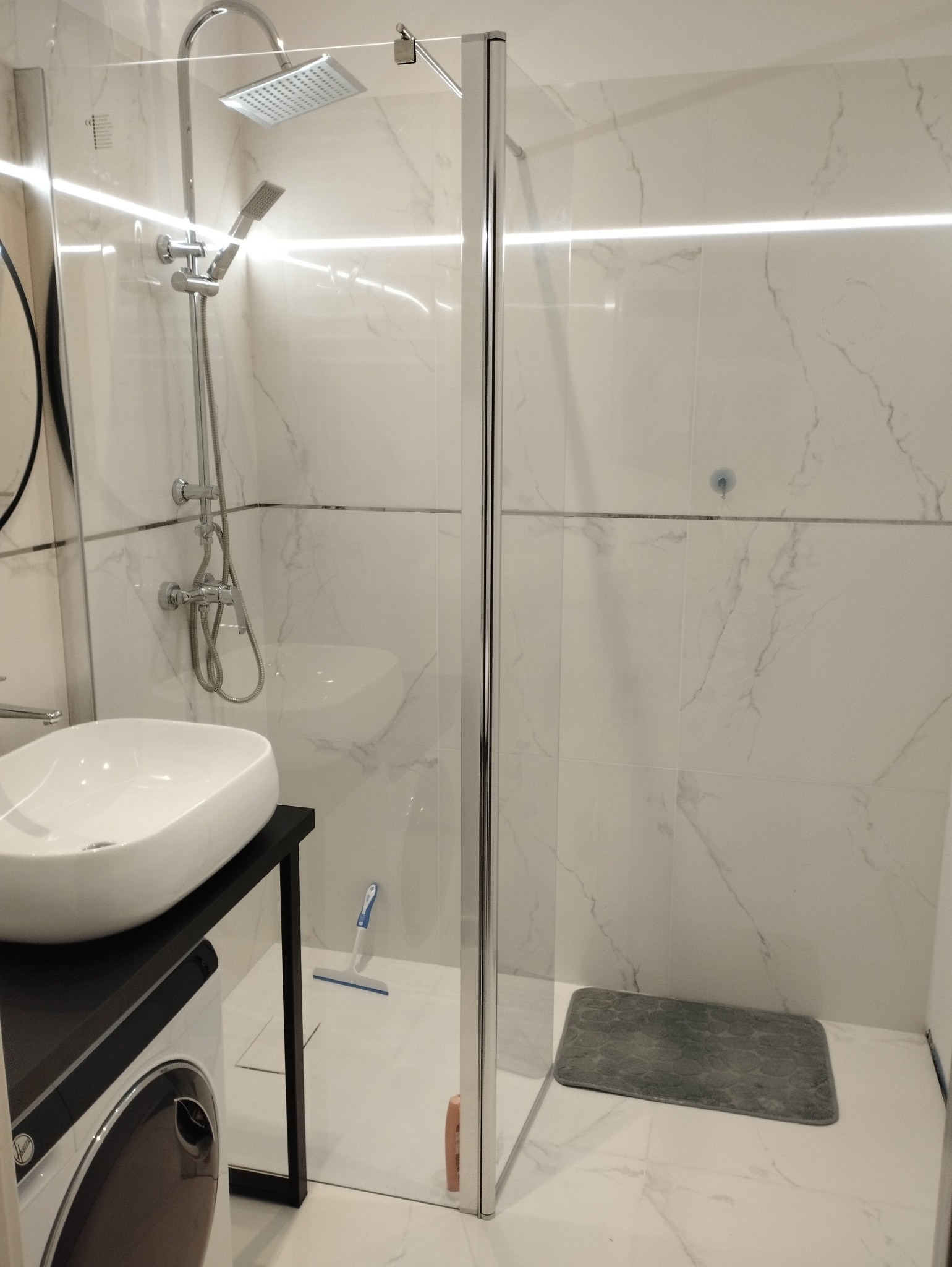 Nowoczesna łazienka z kabiną prysznicową oddzieloną szklaną ścianą, białą umywalką na czarnym blacie i marmurowymi płytkami. Widoczny osprzęt prysznicowy.