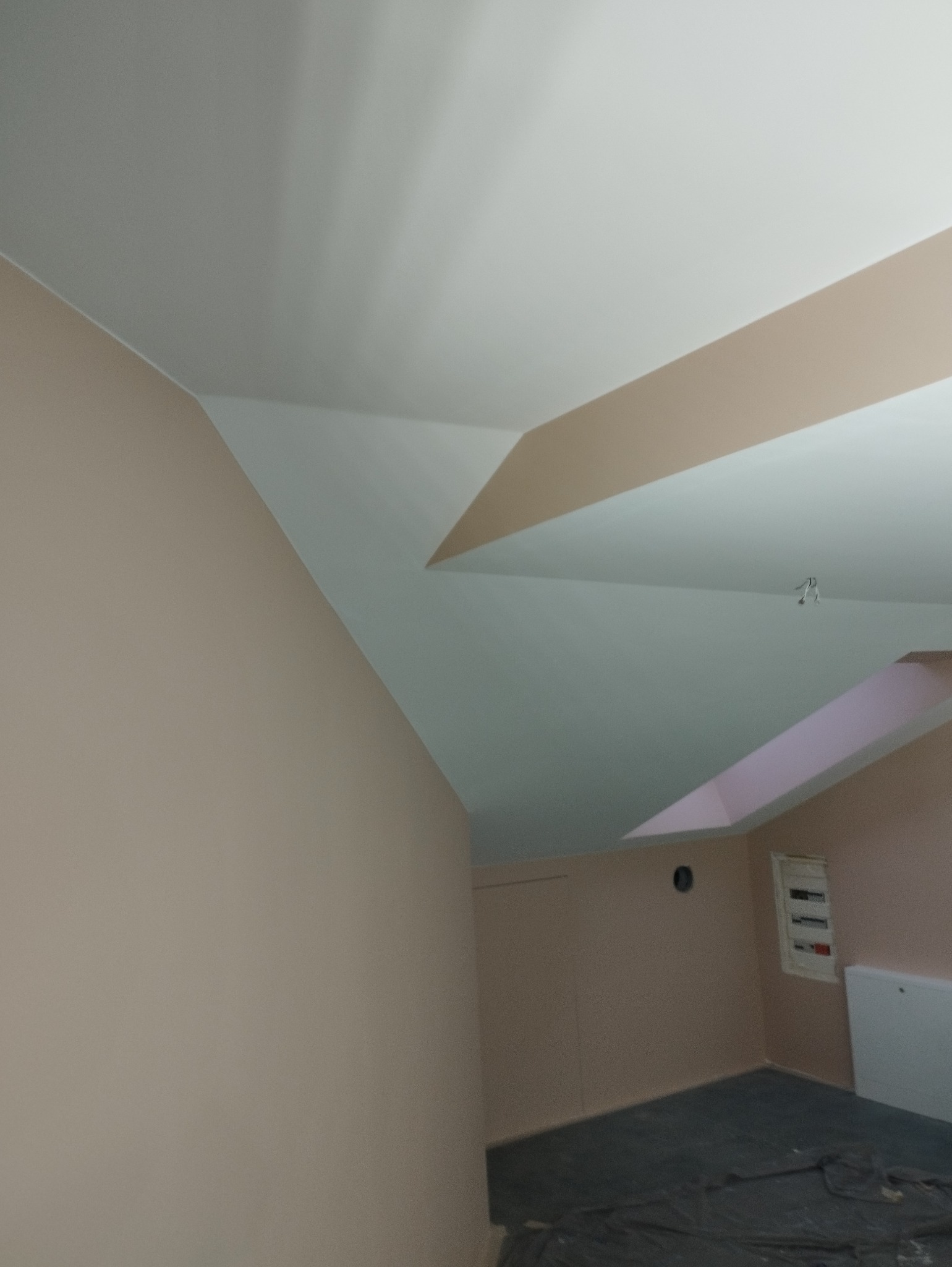 Ściany i sufit w odcieniach beżu i bieli po malowaniu, widoczne załamania sufitu i instalacja elektryczna. Wykończenie wnętrz w pokoju na poddaszu.