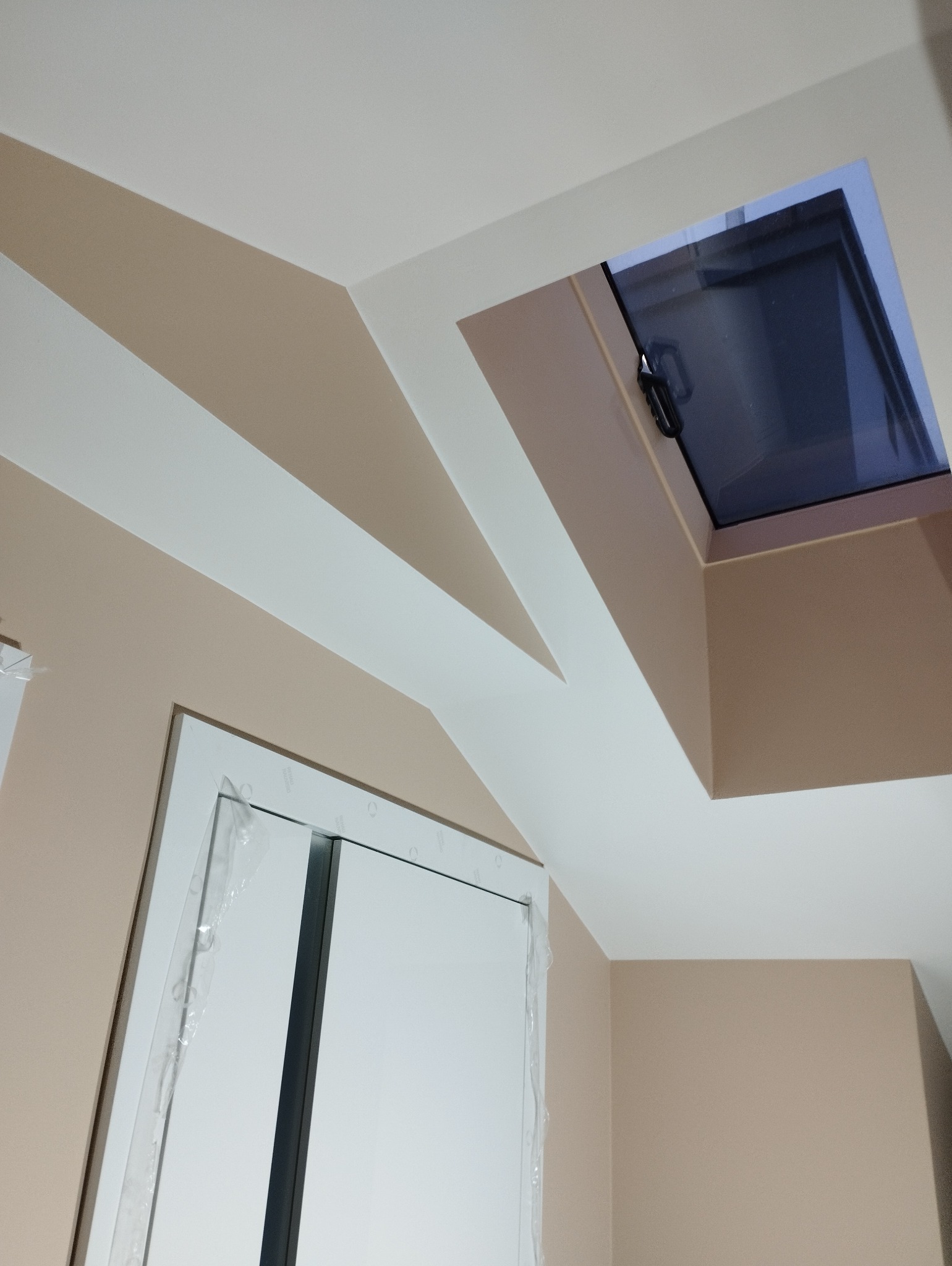 Beżowa ściana z białymi akcentami i oknem dachowym. Widoczne zabezpieczenia na futrynie drzwi. Ukończone malowanie wnętrza.