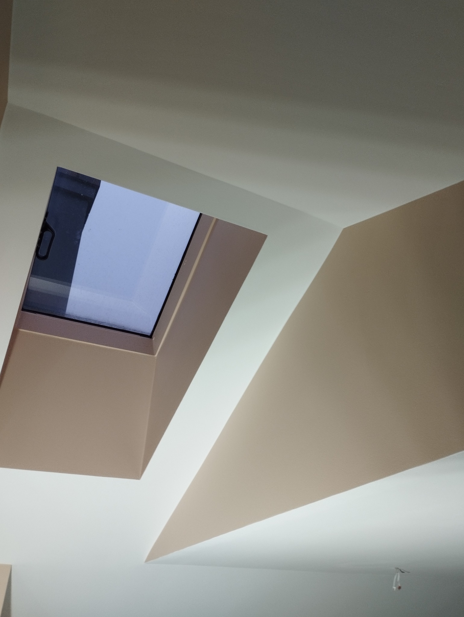 Beżowo-białe ściany i skośny sufit z oknem dachowym. Widok z dołu, podkreślający geometrię wnętrza po malowaniu. Jasne, neutralne barwy.