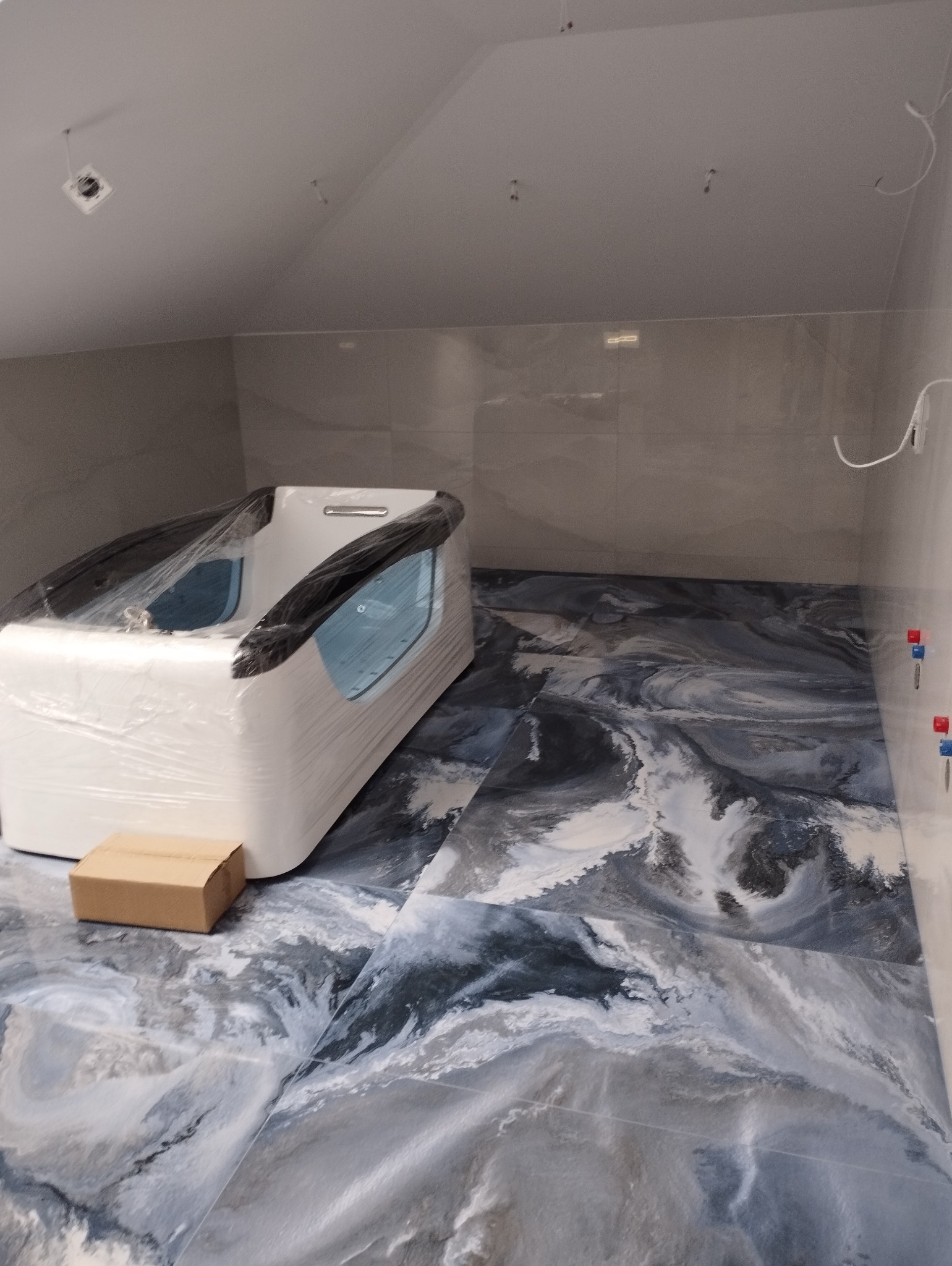 Nowoczesna łazienka w trakcie wykończenia: biała wanna w folii ochronnej na podłodze z żywicy epoksydowej imitującej marmur, szare płytki na ścianach.