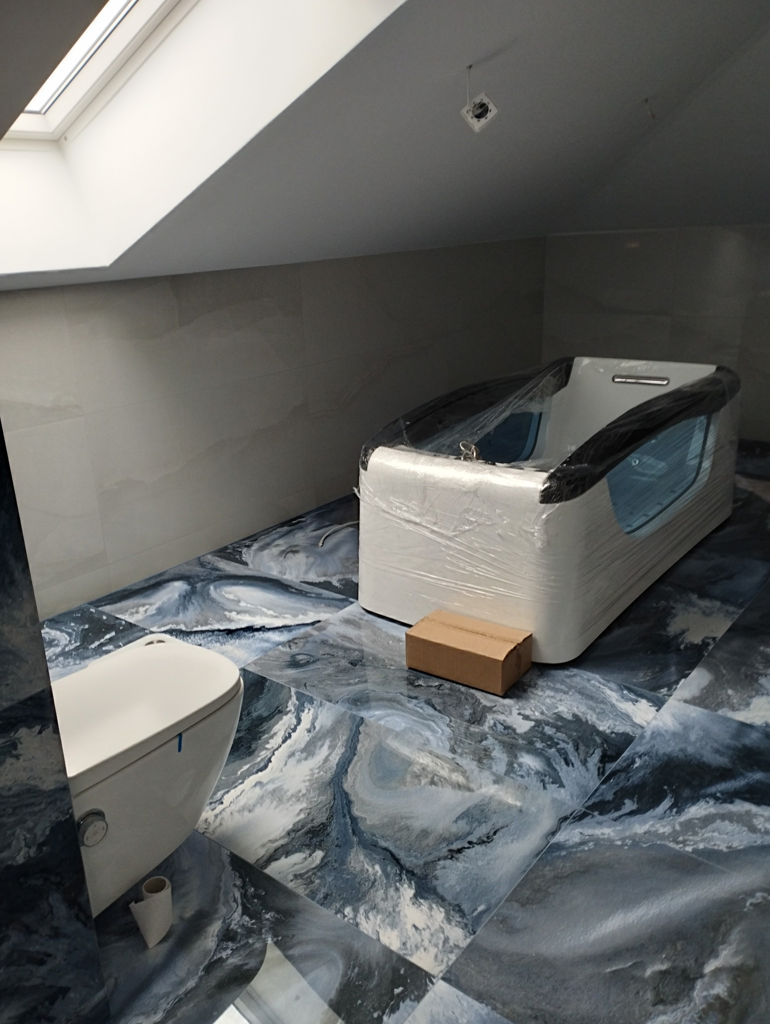 Nowoczesna łazienka na poddaszu z wanną w folii ochronnej, toaletą i podłogą w marmurkowy wzór. Widoczne okno dachowe i surowy punkt oświetleniowy.