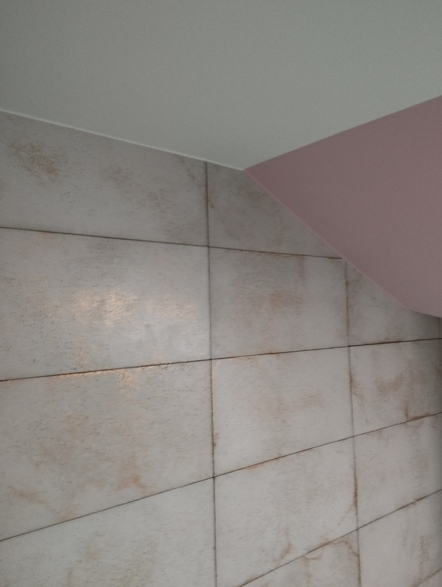 Wykończona ściana w łazience z beżowymi płytkami imitującymi marmur i fragmentem pomalowanego na różowo skośnego sufitu.