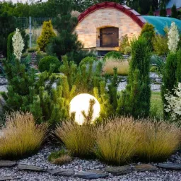 Oświetlony kulisty klosz lampy ogrodowej schowany wśród ozdobnych traw i iglaków na tle ceglanego budynku z zaokrąglonym dachem.