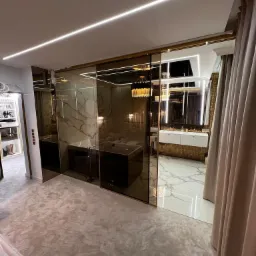 Luksusowa garderoba z wejściem do łazienki oddzielona przesuwnymi taflami szkła w odcieniach brązu, widoczna wanna i marmurowe płytki podłogowe.