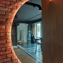 Okrągłe lustro z podświetlaną ramą imitującą czerwoną cegłę, odbijające fragment ekspozycji salonu łazienek z kabiną prysznicową, płytkami i krzesłem barowym.