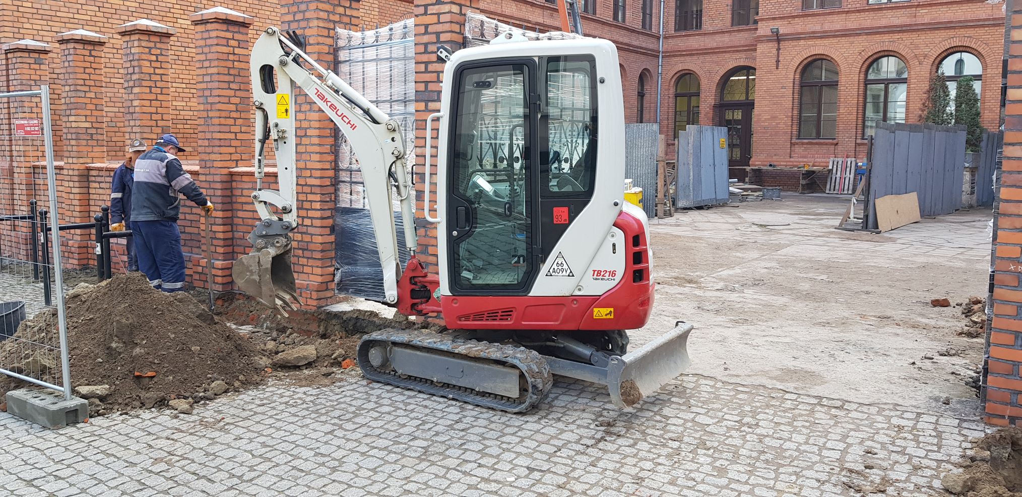 Mini koparka Takeuchi TB216 w trakcie wykonywania wykopu przy ceglanym budynku na brukowanym placu, widoczni dwaj pracownicy w oddali.