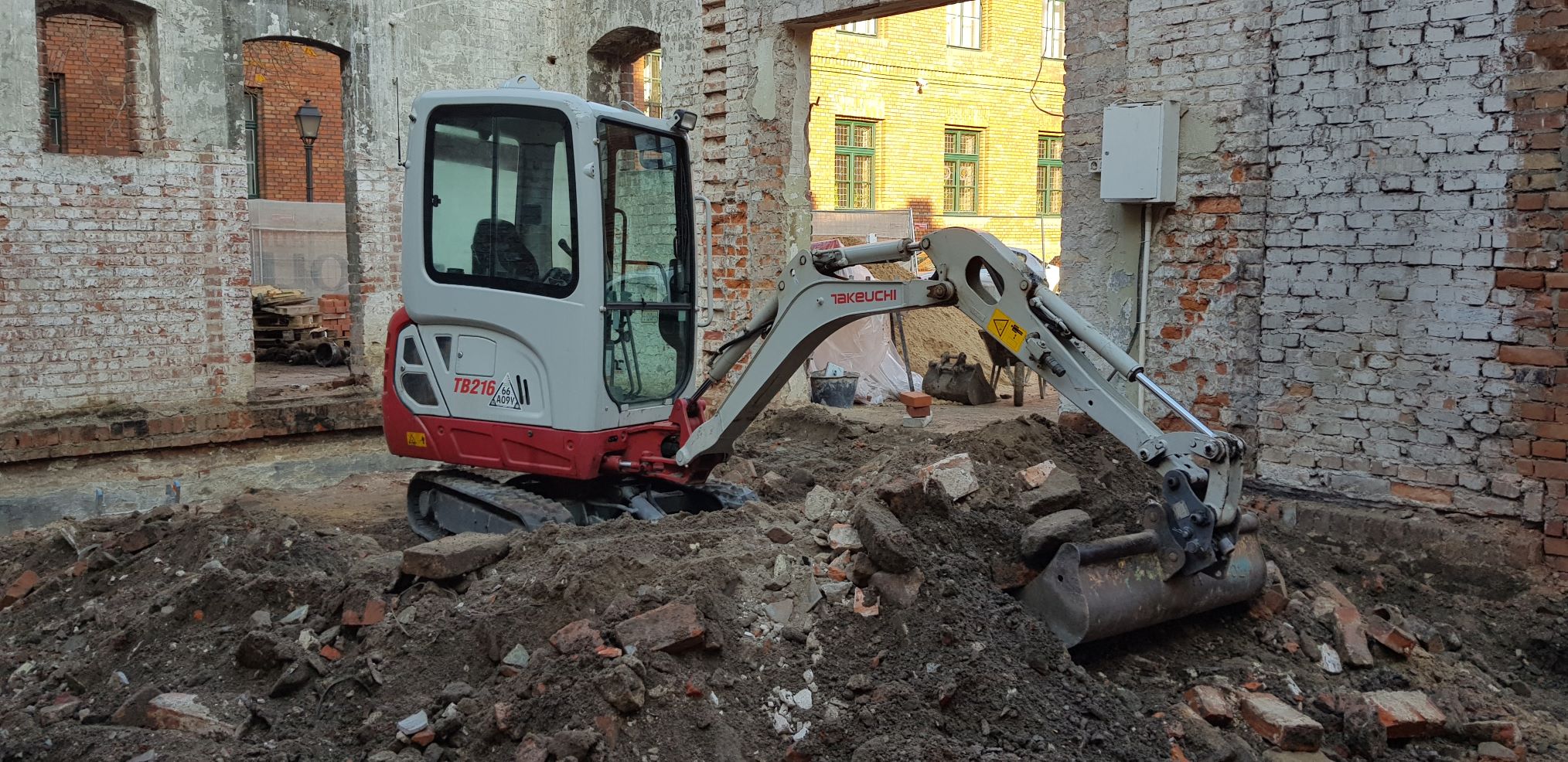 Mini koparka Takeuchi TB216 na placu budowy, z gruzem i ziemią, w tle ceglane ściany i budynek z zielonymi oknami.