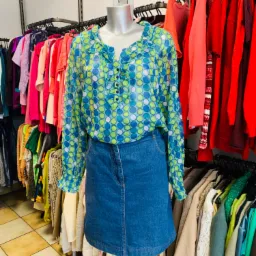 Manekin prezentuje bluzkę w niebiesko-zielone grochy z falbankami i dżinsową spódnicę na tle wieszaków z ubraniami w sklepie z odzieżą używaną.