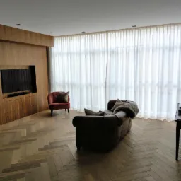 Elegancki salon z parkietem w jodełkę, brązową sofą i kremowymi, transparentnymi zasłonami na całą ścianę okienną. Stonowana kolorystyka i minimalistyczny design.