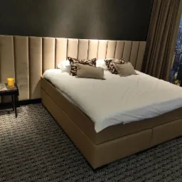 Sypialnia z łóżkiem kontynentalnym, tapicerowanym wezgłowiem w odcieniach beżu i widokiem na miasto za oknem.