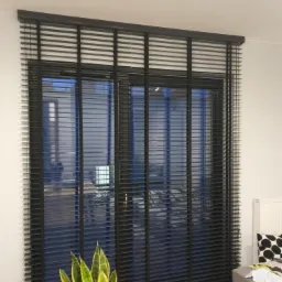 Ciemne, drewniane lamele okienne na dużym oknie tarasowym, z widocznym fragmentem sofy i rośliną doniczkową w minimalistycznym wnętrzu.