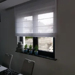 Biała, półprzezroczysta osłona okienna w stylu rzymskim, z widoczną strukturą materiału, zamontowana w nowoczesnym oknie z czarną ramą, na parapecie trzy małe doniczki z zielonymi roślinami...