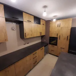 Nowoczesna kuchnia z frontami szafek w odcieniach drewna i czarnymi akcentami, z widocznymi szynami montażowymi i przewodami na ścianie, oraz czarnym blatem i sprzętem AGD.