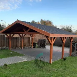 Drewniana konstrukcja z dachem z czarnej dachówki, prawdopodobnie wiata garażowa lub zadaszenie tarasu, widok z zewnątrz, na posesji z kostką brukową i fragmentem trawnika.