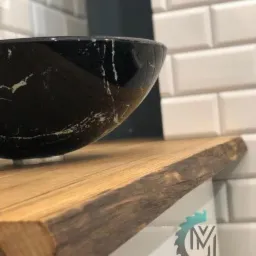 Czarna umywalka marmurkowa na blacie z jasnego drewna o naturalnej krawędzi, tło z białej kafelkowej ściany typu metro, widoczny logo firmy na blacie.