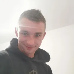 Uśmiechnięty mężczyzna w czarnej bluzie z kapturem i nadrukiem lwa z koroną, patrzący w górę na jasne białe tło.