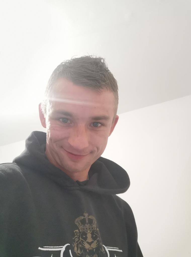 Uśmiechnięty mężczyzna w czarnej bluzie z kapturem i nadrukiem lwa z koroną, patrzący w górę na jasne białe tło.