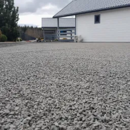 Szara, kamienna nawierzchnia podjazdu przed nowoczesnym domem z białymi ścianami i szarą dachówką, widok z niskiej perspektywy.