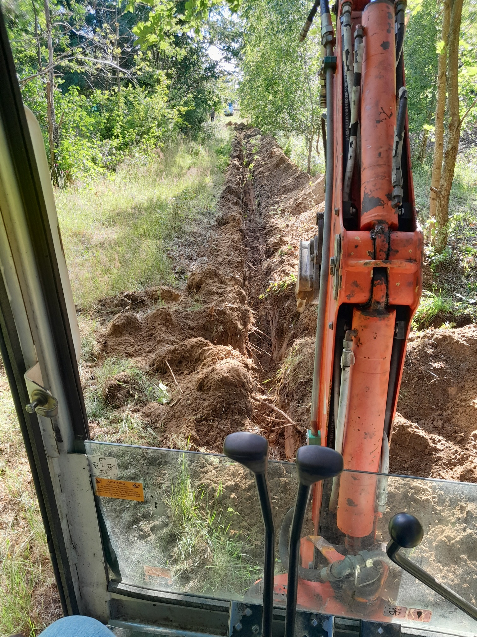 Widok z kabiny pomarańczowej koparki na wykopany rów w ziemi, otoczony zielenią i drzewami. Rów ciągnie się w górę zbocza, a w oddali widoczny jest inny pojazd.