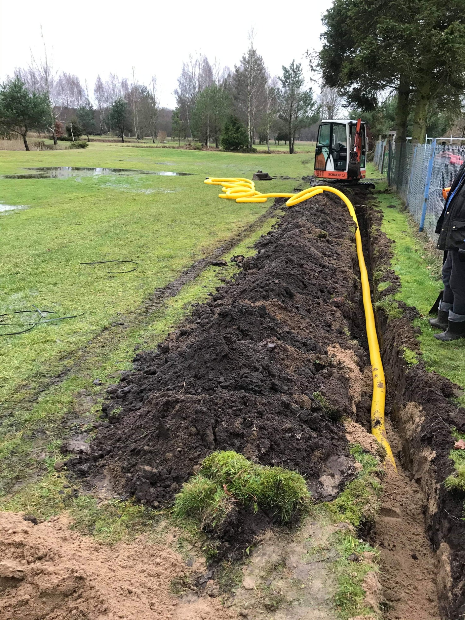 Żółta rura ułożona w wykopie na terenie podmokłym, z widocznym mini-koparką i osobą w roboczym ubraniu w tle.