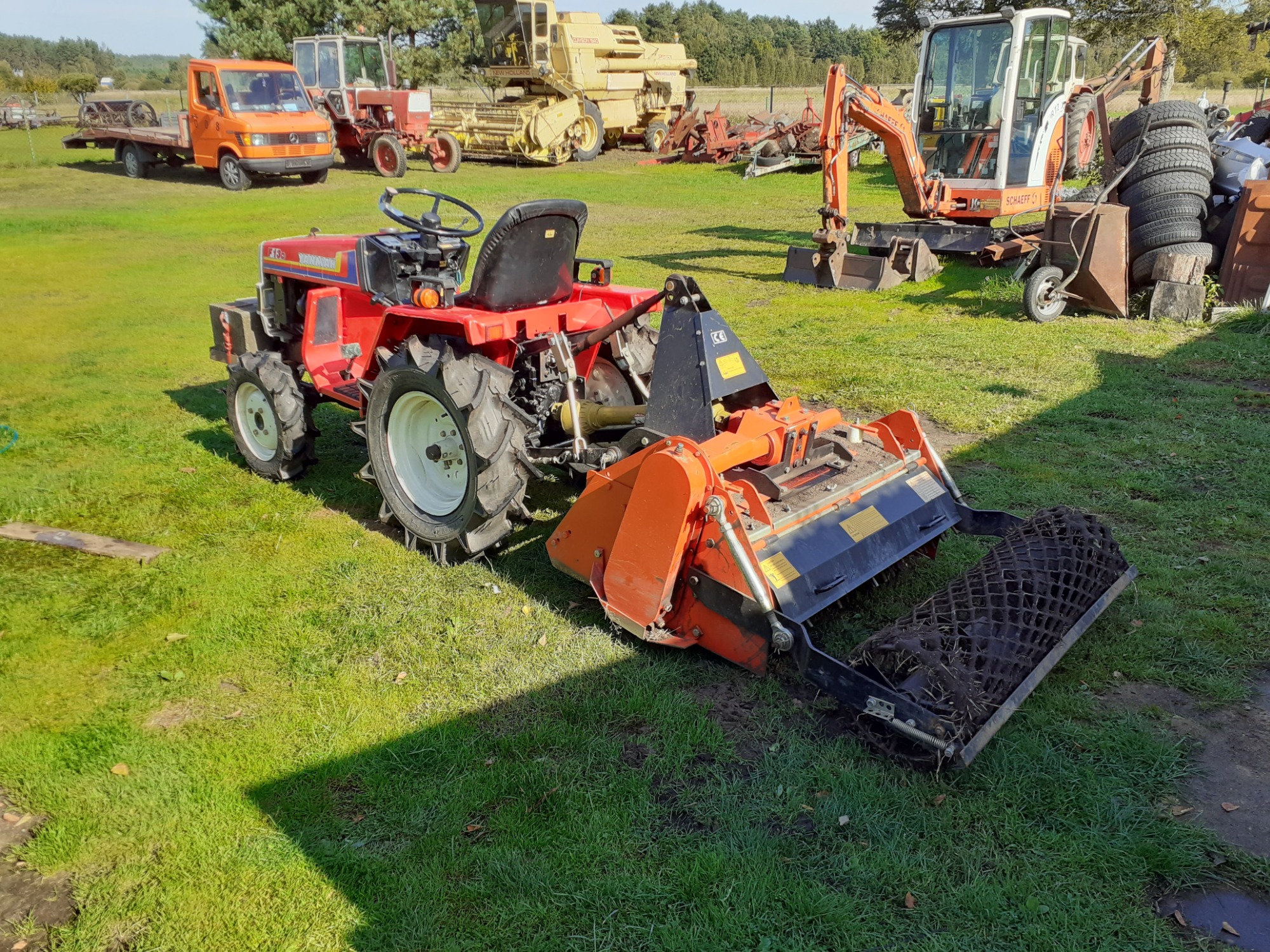 Czerwony miniciągnik z glebogryzarką na trawiastym terenie, w tle inne maszyny rolnicze i budowlane, w tym pomarańczowa laweta i koparka.