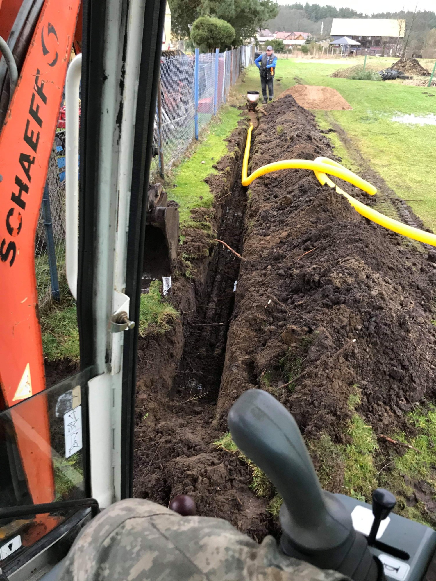 Wąski wykop w ziemi z ułożoną żółtą rurą, widziany z kabiny pomarańczowej koparki Schaeff, operator w odzieży moro, w tle ogrodzenie z siatki i pracownik.