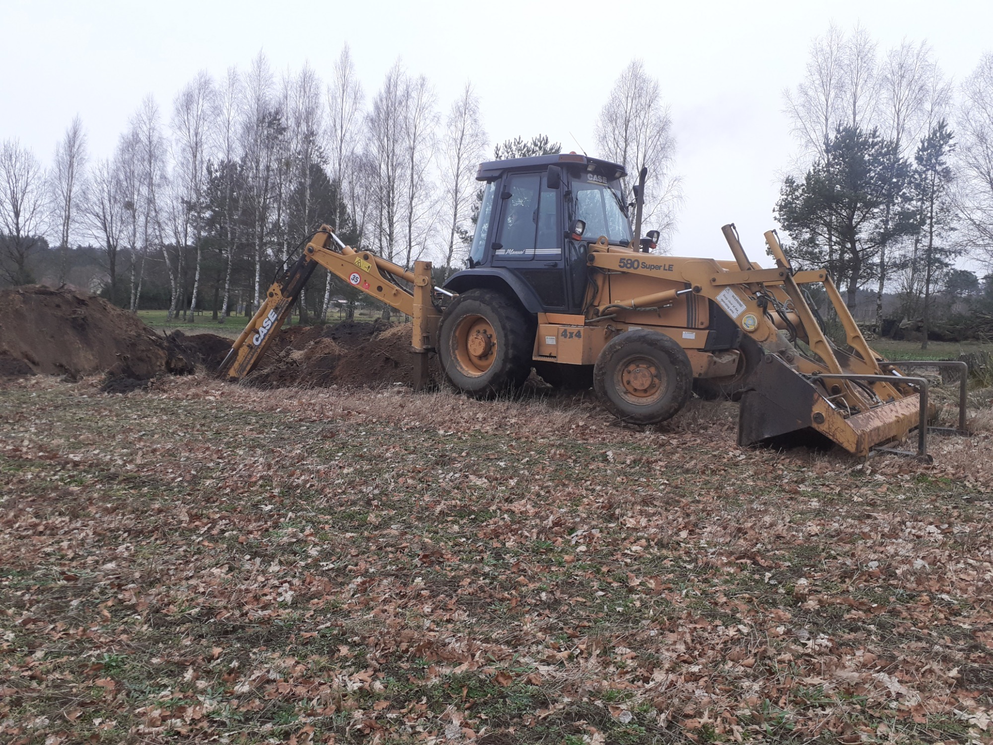 Żółta koparko-ładowarka Case 580 Super LE, z widocznym napisem 4x4, podczas prac ziemnych na polu pokrytym liśćmi, z drzewami w tle.
