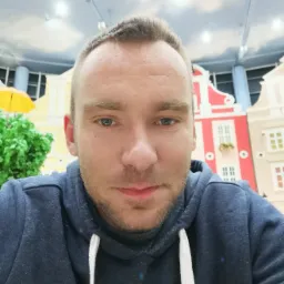 Portret mężczyzny w bluzie na tle dekoracji imitującej kolorowe kamienice i błękitne niebo. Widoczny fragment żółtego parasola i zieleni.