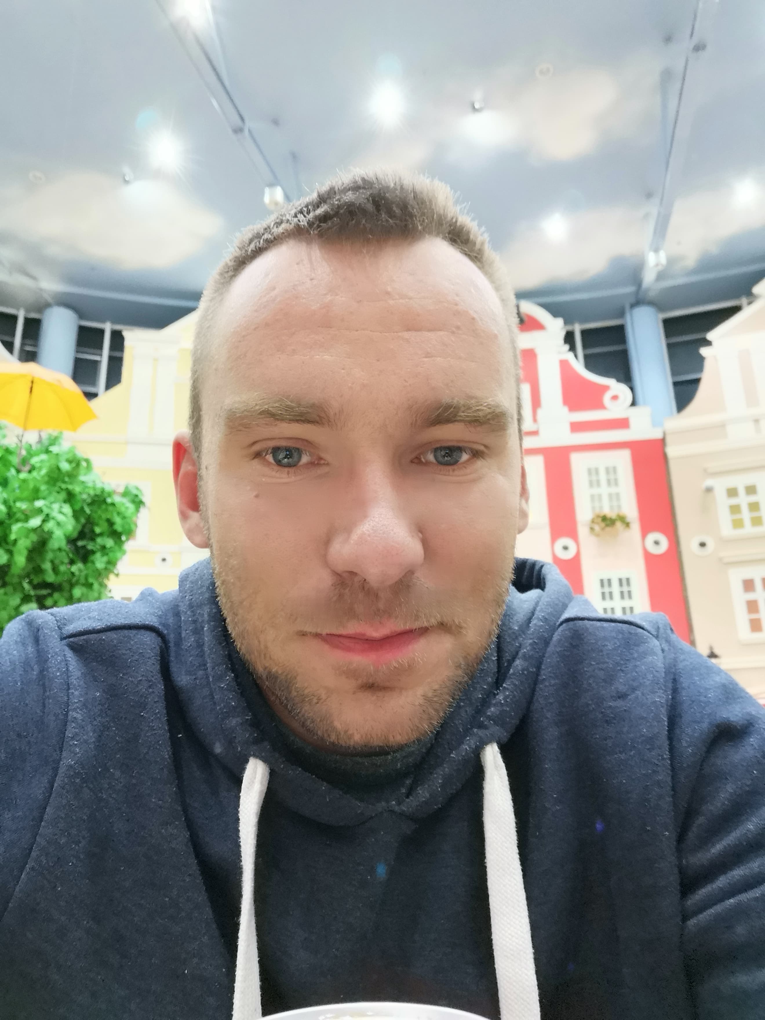 Portret mężczyzny w bluzie na tle dekoracji imitującej kolorowe kamienice i błękitne niebo. Widoczny fragment żółtego parasola i zieleni.