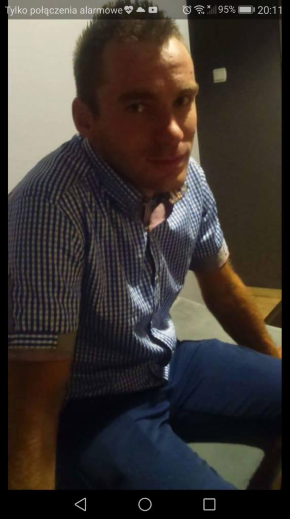 Portret mężczyzny w niebieskiej koszuli w kratę i spodniach, wykonany telefonem komórkowym z widocznym paskiem stanu.