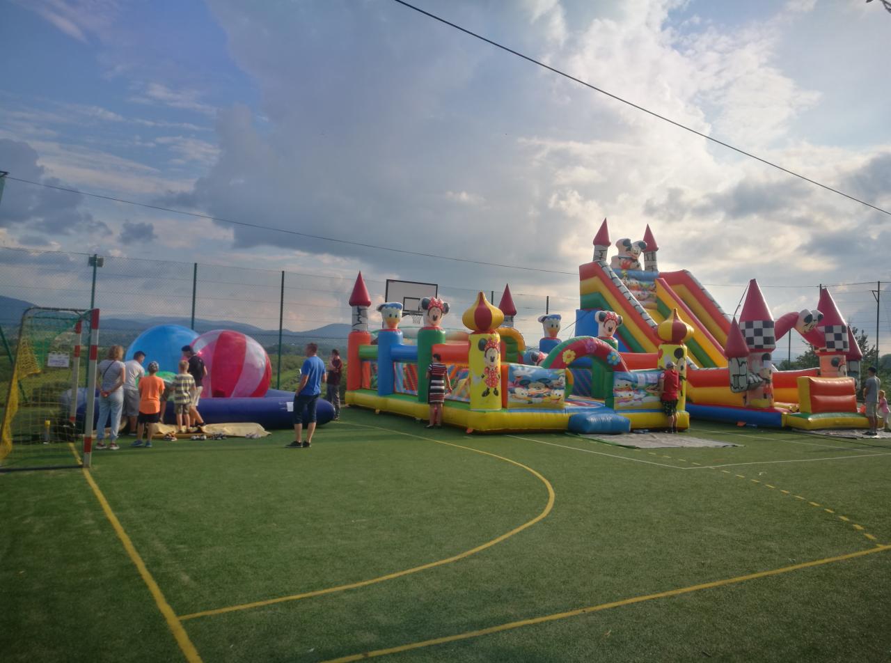 Kolorowe zamki dmuchane z motywami z bajek rozstawione na zielonym boisku sportowym, w tle widoczni ludzie i zalesione wzgórza pod pochmurnym niebem.