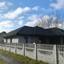 Nowoczesny dom jednorodzinny z ciemną dachówką ceramiczną i betonowym ogrodzeniem, widok z zewnątrz w słoneczny dzień.