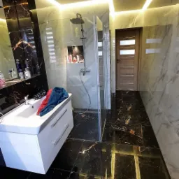 Nowoczesna łazienka z czarnymi marmurowymi płytkami podłogowymi i ściennymi, białą umywalką z szafką, szklaną kabiną prysznicową i oświetleniem LED na suficie.