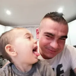 Uśmiechnięty mężczyzna z irokezem i mały chłopiec wyciągający język na tle białego sufitu, w kadrze od dołu.