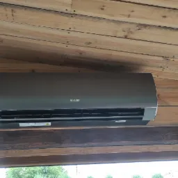 Szary klimatyzator marki Sinclair zamontowany na drewnianej ścianie wewnątrz pomieszczenia z drewnianym sufitem i ramą okienną.