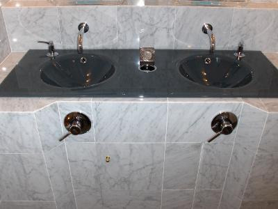 Podwójna umywalka w łazience wykończonej marmurowymi płytkami, z czarnym blatem i chromowanymi bateriami, oraz dozownikiem mydła i szklanką na szczoteczki.