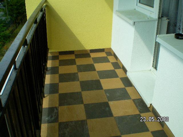 Balkon z szachownicą z płytek w odcieniach brązu i czerni, widok z góry, żółta ściana w tle, czarna barierka po lewej stronie.