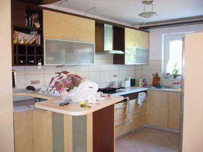 Kuchnia w trakcie remontu: widoczne szafki w odcieniach beżu i brązu, na blacie leżą narzędzia i kwiaty w plastikowej torbie, ściany wyłożone jasnymi płytkami.