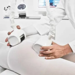 ICOONE: Osoba w białym kombinezonie poddawana zabiegowi na ciało z użyciem specjalistycznego urządzenia z dwiema głowicami masującymi, widoczny panel sterowania.