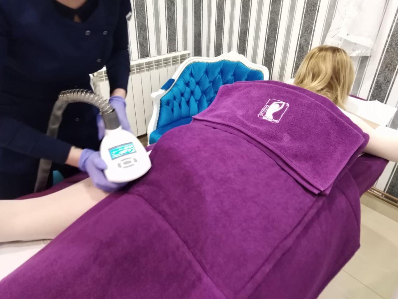 Klientka leży na łóżku przykryta fioletowym ręcznikiem z logo studia, podczas zabiegu urządzeniem do modelowania sylwetki przez osobę w niebieskim uniformie i fioletowych rękawiczkach.