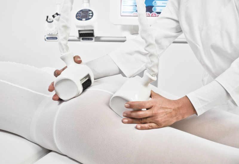 ICOONE: Osoba w białym kombinezonie poddawana zabiegowi na ciało z użyciem specjalistycznego urządzenia z dwiema głowicami masującymi, widoczny panel sterowania.