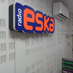 Podświetlany szyld z logo 'Radio Eska' w pomarańczowo-niebieskich barwach, zamontowany na białej ścianie z perforowanymi otworami, obok klimatyzatora i lampki podłogowej.