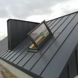 Szary dach z blachy trapezowej z oknem dachowym Fakro i kominem wentylacyjnym w pochmurny dzień.