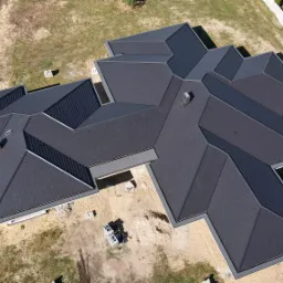 Widok z lotu ptaka na nowoczesny dom z ciemnoszarym, wielospadowym dachem z widocznymi kominkami wentylacyjnymi i obróbkami blacharskimi, częściowo wykończony, z materiałami budowlanymi na działce.