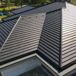 Nowoczesny, ciemny dach z dachówki ceramicznej widziany z góry, z widocznymi elementami wykończeniowymi i fragmentem budynku w stanie surowym otwartym.