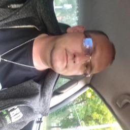 Adam Przyborowski - Mężczyzna w okularach, w samochodzie, ubrany w bluzę z kapturem i czarną koszulkę z łańcuszkiem. Widok z boku, oświetlenie naturalne.