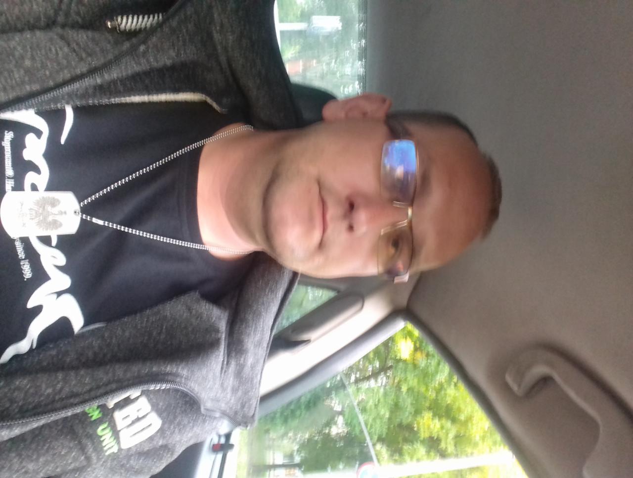 Mężczyzna w okularach, w samochodzie, ubrany w bluzę z kapturem i czarną koszulkę z łańcuszkiem. Widok z boku, oświetlenie naturalne.